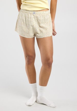 Personne portant un short jaune à fleurs, un haut jaune clair et des chaussettes blanches, debout devant un fond gris clair uni.