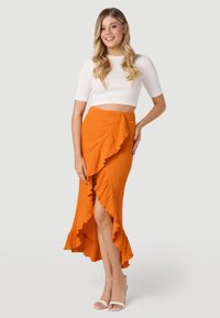 Oranžová midi sukňa s volánmi a vysokým rozparkom, textúrovaná látka a priliehavý pás; skombinovaná s bielym skráteným topom a podpätkami.