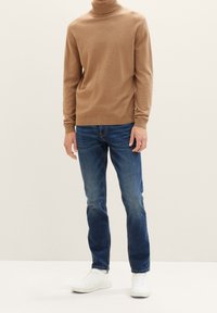 Suéter de cuello alto marrón hecho de una tela suave, combinado con vaqueros azules y zapatillas blancas. Presenta puños acanalados y un diseño ajustado.