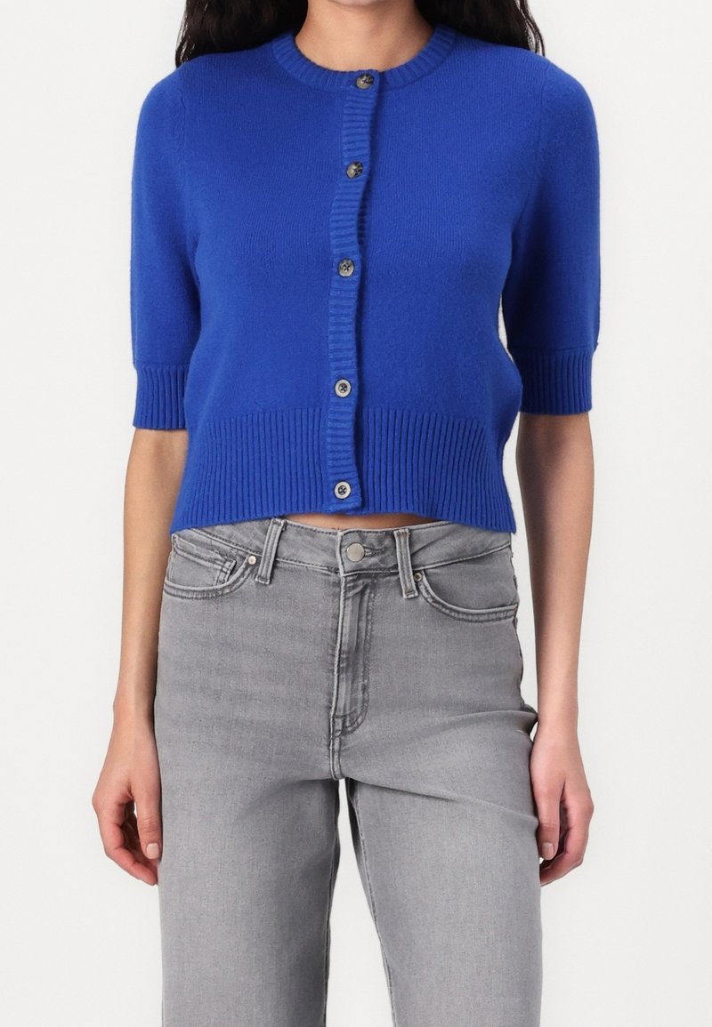 Femme portant un cardigan bleu vif à manches courtes boutonné, associé à un jean gris clair taille haute.