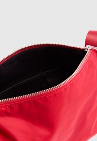 Κόκκινη τσάντα από νάιλον με μαύρο εσωτερικό, μεγάλο φερμουάρ και υφασμένη ράβδο. Η τσάντα έχει λείο, γυαλιστερό εξωτερικό και μεταλλική λεπτομέρεια φερμουάρ.