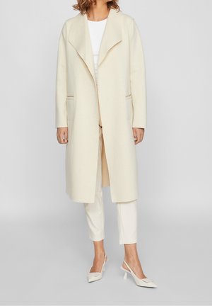 Vrouw die een lange crèmekleurige jas, witte top, witte cropped broek en witte slingback hakken draagt, staat tegen een effen achtergrond.