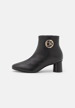 Bottines noires en cuir lisse, avec un bout pointu et un talon cylindrique. Un accent circulaire en or avec le logo est affiché de manière proéminente.