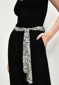 Ceinture en tissu à motif léopard nouée à la taille, affichant un motif noir et blanc. Portée par-dessus un pantalon taille haute noir, avec un haut texturé.