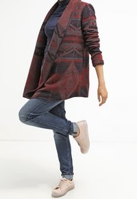 Personne portant une veste à motifs rouge et noir, un haut bleu marine, un jean bleu et des baskets rose clair, en équilibre sur une jambe, l'autre jambe fléchie.