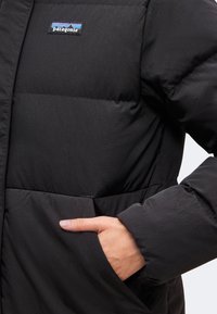 Χέρι στην τσέπη μαύρου μονωμένου μπουφάν Patagonia με ορατό λογότυπο στο στήθος πάνω σε απλό φόντο.