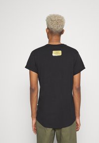 T-shirt preto de manga curta com bainha curvada, apresentando um rótulo amarelo na parte de trás com texto. Feito de um tecido macio e leve.