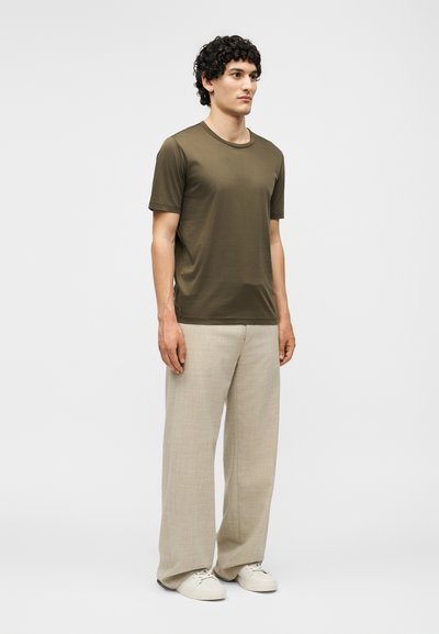 Νεαρός άντρας που στέκεται προς τα εμπρός, φοράει ένα ελιάς πράσινο t-shirt, μπεζ παντελόνι με φαρδιά πόδια και λευκά αθλητικά παπούτσια σε απλό φόντο.