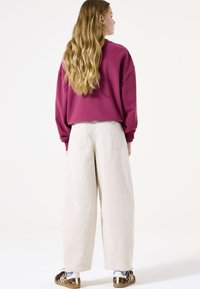 Sudadera de gran tamaño color burdeos con hombros caídos, combinada con pantalones anchos de color beige claro. Pies en zapatos de lona con patrones sobre un fondo liso.