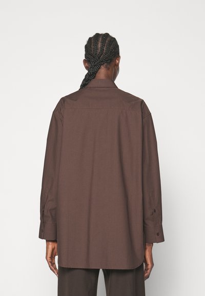Cămașă oversized de culoare maro, dintr-un material fin, cu guler, mâneci lungi și manșete cu nasturi; prezintă un tiv drept și detaliu la spate.