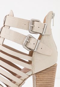 Sandales à talons hauts beige avec des lanières, texture crocodile, boucles métalliques et talon à texture bois. Conçues avec des détails ajourés.