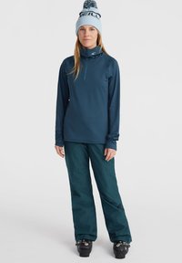 Maglia a maniche lunghe blu navy con colletto alto e mezza cerniera, abbinata a pantaloni larghi di un verde blu scuro. Stivali da sci neri con fibbie regolabili.