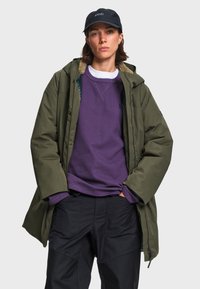Femme portant un parka à capuche vert foncé, un pull violet, un pantalon noir et une casquette noire, debout devant un fond clair uni.