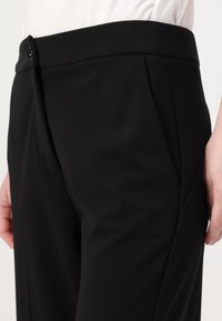 Pantaloni neri sartoriali con una texture liscia, vita con chiusura a bottone e tasche laterali, che mostrano linee pulite e una forma aderente.