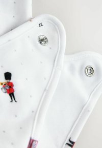 Vitt tyg med mjuk textur prytt med små tryckta mönster. Inkluderar ett dekorativt motiv av en figur i röd kappa som håller en trumpet.