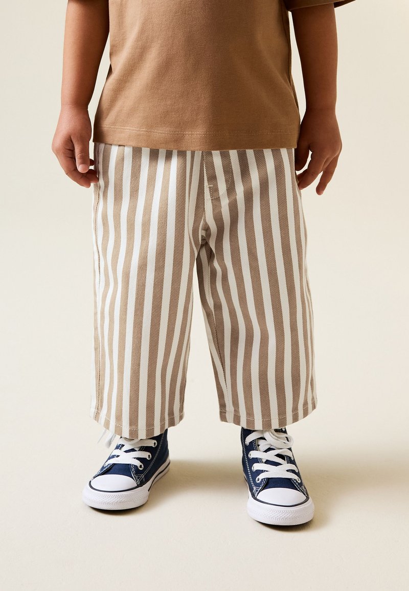 Barn klädd i brun tröja, beige och vita vertikalt randiga byxor samt marinblå snörskor stående på ett ljust golv.