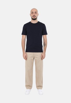 Uomo con barba e tatuaggio in piedi di fronte, indossa una maglietta nera, pantaloni beige e scarpe da ginnastica bianche su sfondo chiaro semplice.