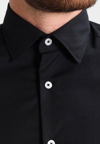 Camicia nera con bottone realizzata in tessuto liscio. Presenta un colletto classico, design semplice e bottoni bianchi con un centro nero.
