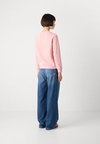 Persoon met kort donker haar, gekleed in een roze longsleeve en losse blauwe jeans, staande tegenover een witte muur.