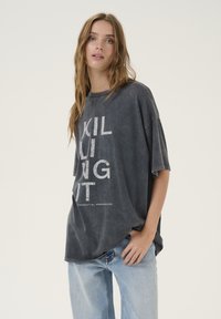 Tmavohnedé oversized tričko s krátkymi rukávmi a vyblednutou textúrou. Na prednej strane je vytlačený biely text "KILLING IT" a "ESSENTIAL WARDROBE".