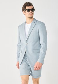 Completo sartorial azzurro chiaro con un solo bottone, abbinato a pantaloni corti coordinati. Il tessuto sembra testurizzato. Il modello indossa occhiali da sole scuri.