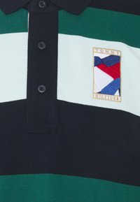 Polo en coton avec des rayures horizontales vertes, blanches et marine, patte de boutonnage marine, et logo brodé de drapeau Tommy Hilfiger sur la poitrine.