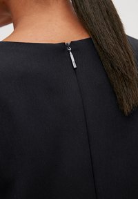 Schwarzes Kleid mit einem verborgenen Reißverschluss auf der Rückseite und silbernem Zipper, gefertigt aus strukturiertem Stoff, der klare Linien und ein minimalistisches Design zeigt.