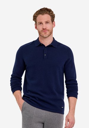 Man with light brown hair and beard wearing a navy blue long-sleeve polo and gray pants with one hand in pocket.