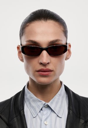 Osoba s uhlazenými vlasy, která má na sobě úzké tmavé brýle, světlou pruhovanou košili s knoflíky a černou koženou bundu na bílém pozadí.