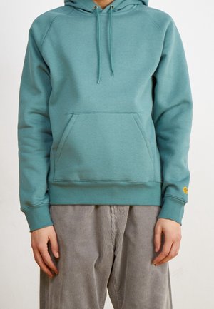 Personne portant un sweat à capuche bleu sarcelle avec une poche avant et un pantalon en velours côtelé gris, debout devant un fond clair uni.