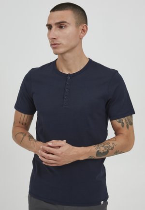 Mørkeblå kortermet henley-skjorte med rund hals og fire-knaps placket, laget av mykt materiale. Viser synlige tatoveringer på armene.