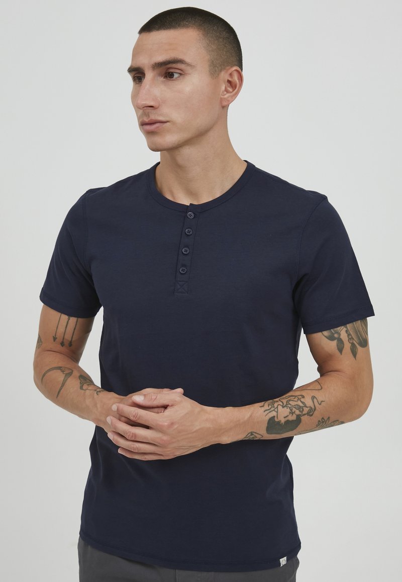 Mørkeblå kortermet henley-skjorte med rund hals og fire-knaps placket, laget av mykt materiale. Viser synlige tatoveringer på armene.
