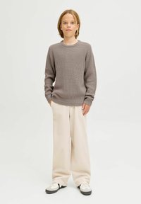 Grauer Strickpullover mit gerippter Textur, kombiniert mit lockeren, cremefarbenen Hosen und Schwarz-Weiß-Sneakern. Weißer Hintergrund.