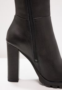 Botte cheville en cuir noir avec talon bloc, fermeture éclair latérale et semelle texturée sur un fond clair.