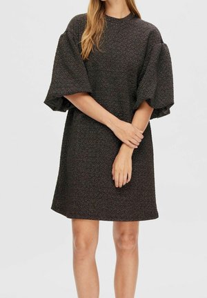 Kvinde iført en mørkegrå tekstureret kort kjole med brede, puffede ærmer, stående med hænderne løst krydset foran sig.