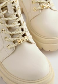 Witte leren enkelboots met een getextureerde afwerking, gouden accenten, gevlochten veters en strikdetail, met een dikke rubberen zool.