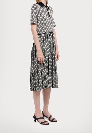 Femme debout portant une robe à manches courtes à motif noir et blanc avec une jupe plissée et des sandales noires à talons sur un fond uni.