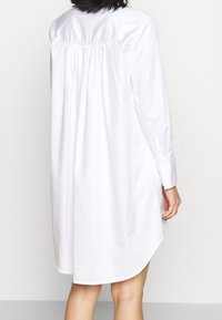 Persona che indossa un abito camicia bianco, lungo e a maniche lunghe, visto da dietro su uno sfondo chiaro e semplice.