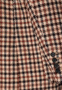 Tissu en mélange de laine avec un motif à carreaux beige comprenant des nuances de marron, bleu marine et rouge ; six boutons noirs alignés sur le côté droit.