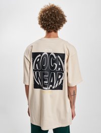 Bežs pārāk liels T-kreklis ar lielu melnbaltu "ROCA WEAR" grafiku aizmugurē, ar "EST. 1999" dzeltenā krāsā. Kokvilnas audums.