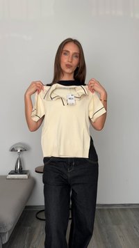 Vrouw die een crème kortmouwtrui met zwarte afwerking en een prijskaartje vasthoudt, staande in een minimalistische kamer met een lamp en boeken.