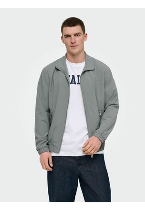 Ung mann iført en lys grå jakke med glidelås over en hvit skjorte med marineblå tekst og mørkeblå jeans, stående foran en ensfarget bakgrunn.