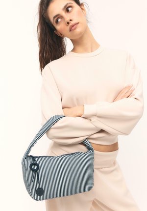 Femme aux cheveux bruns attachés en queue de cheval, vêtue de vêtements beige clair, tenant un petit sac à main rayé bleu et blanc avec une fermeture éclair et un charm décoratif.