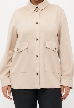 Beżowa kurtka-koszula z dwoma klapowymi kieszeniami, noszona na czarnych spodniach, pokazana od ramion do bioder.