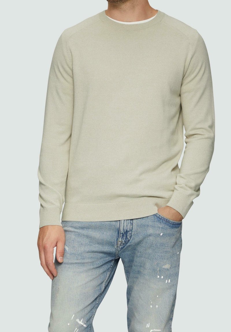 Hellgrüner Strickpullover mit rundem Ausschnitt und gerippten Bündchen, kombiniert mit zerfetzten blauen Jeans. Weiche Textur und figurbetontes Design.