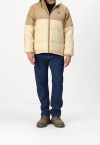 Puffy beige and light tan jacket with a front zipper, ribbed cuffs, and a small patch logo on the left chest. Worn with blue jeans and brown shoes.