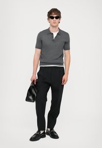 Grå kortærmet polo shirt, sorte skræddersyede bukser, sorte lædersko og en sort taske. Mand iført solbriller, stående foran en hvid baggrund.