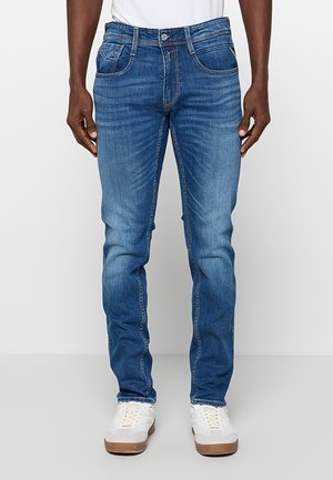 Homme portant un jean slim bleu moyen avec des détails de whiskering, associé à des baskets blanches à semelles en gomme, debout devant un fond uni.