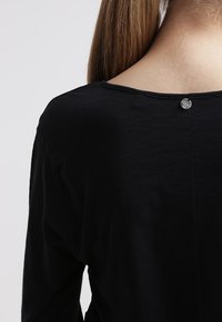 Schwarzes Langarmshirt mit Rundhalsausschnitt und einem kleinen Metallknopfverschluss auf der Rückseite. Weicher, glatter Stoff mit leichtem Glanz.