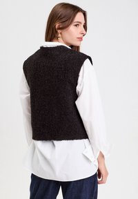 Schwarze, strukturierte Weste über einem weißen Langarmshirt; hat einen verkürzten Schnitt und weite Armausschnitte, die ein sauberes Design zur Geltung bringen.